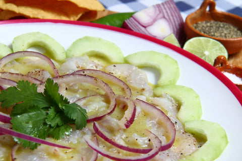 Aguachile de camarón con cebolla morada, pepino y cilantro en un plato blanco, con tostadas, limones y pimienta al fondo