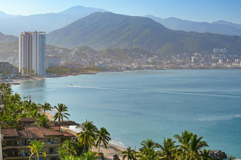 Vista elevada de Puerto Vallarta: playa con palmeras y techos de teja, una lancha cruzando la bahía y la ciudad al pie de la sierra verde